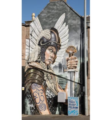 A large outdoor wall mural featuring a blonde female warrior or Valkyrie wearing a winged helmet, armor, and arm tattoos, holding an upright sword against a misty landscape backdrop.