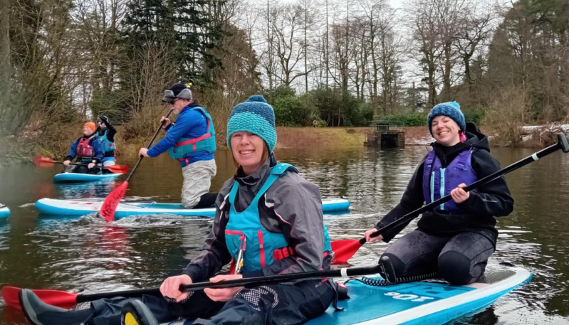 Participants wearing buoyancy aids paddle stand-up paddleboards across a calm loch, with trees and parkland visible along the shoreline.