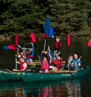 Group of children and an instructor in two open canoes on a loch, holding colourful paddles aloft during an outdoor water activity.