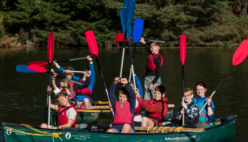 Group of children and an instructor in two open canoes on a loch, holding colourful paddles aloft during an outdoor water activity.