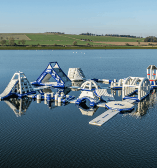 Aerial view of a large inflatable aqua park floating on a loch, featuring climbing frames, slides, balance obstacles and platforms surrounded by open countryside.