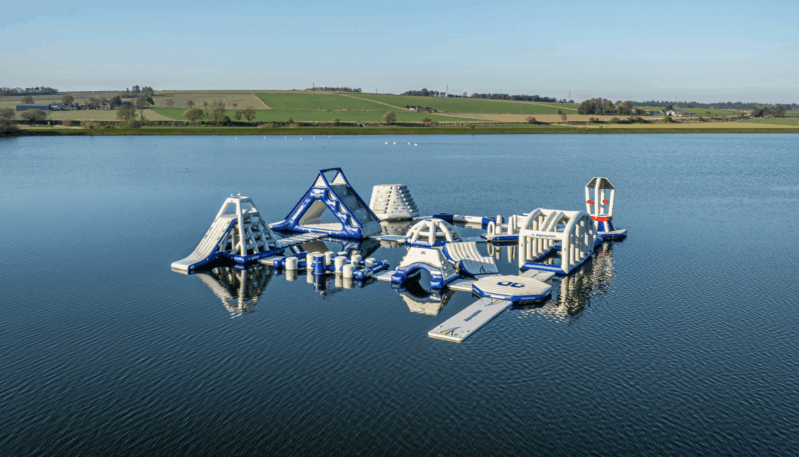 Aerial view of a large inflatable aqua park floating on a loch, featuring climbing frames, slides, balance obstacles and platforms surrounded by open countryside.