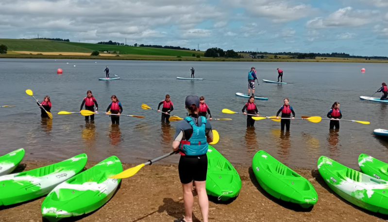 Outdoor watersports session at a loch, with participants wearing buoyancy aids receiving kayak and paddleboard instruction on the shoreline, while others practise paddleboarding on the water behind them.