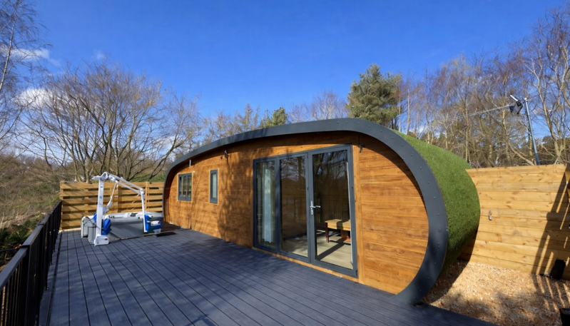 A modern, curved wooden cabin with a grass-covered roof sits on a dark grey deck. The "escape pod" features large glass doors and small windows, set against a backdrop of bare trees and a clear blue sky. A privacy fence and a white accessibility lift are visible on the deck.