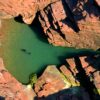 An aerial, top-down view of a secluded, turquoise sea cave pool nestled within jagged, reddish-brown cliffs. Two people swim in the clear water, while several colorful kayaks and a small group of people are gathered on a tiny pebble beach at the water's edge.