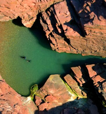 An aerial, top-down view of a secluded, turquoise sea cave pool nestled within jagged, reddish-brown cliffs. Two people swim in the clear water, while several colorful kayaks and a small group of people are gathered on a tiny pebble beach at the water's edge.