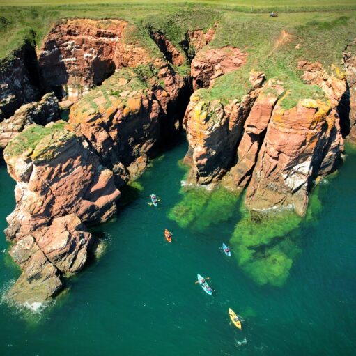 A high-angle aerial shot of several bright kayaks paddling through vibrant turquoise water along a rugged coastline. The scene features dramatic, towering red sandstone cliffs and sea stacks topped with green grass under a clear, sunny sky.