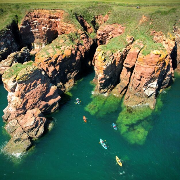 A high-angle aerial shot of several bright kayaks paddling through vibrant turquoise water along a rugged coastline. The scene features dramatic, towering red sandstone cliffs and sea stacks topped with green grass under a clear, sunny sky.
