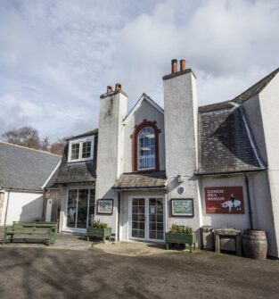 The exterior of the Glenesk Folk Museum, a charming, light-colored stone and stucco building with two prominent chimneys and a central arched window. A red and white sign for the museum is visible on the right. A small outdoor seating area with picnic benches is on the left, under a partly cloudy sky.