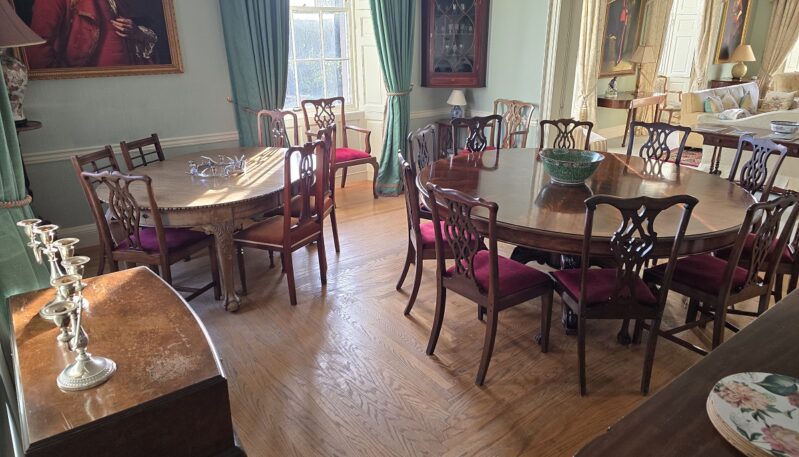 A grand dining room with two large, polished mahogany tables and matching velvet-cushioned chairs. The room is decorated with period portraits, green curtains, and traditional wooden cabinetry.