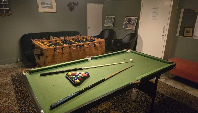 A leisure room featuring a pool table with cues and balls, a table football (foosball) game, and dark leather armchairs. The walls are a muted green, accented by framed artwork and a decorative rug.