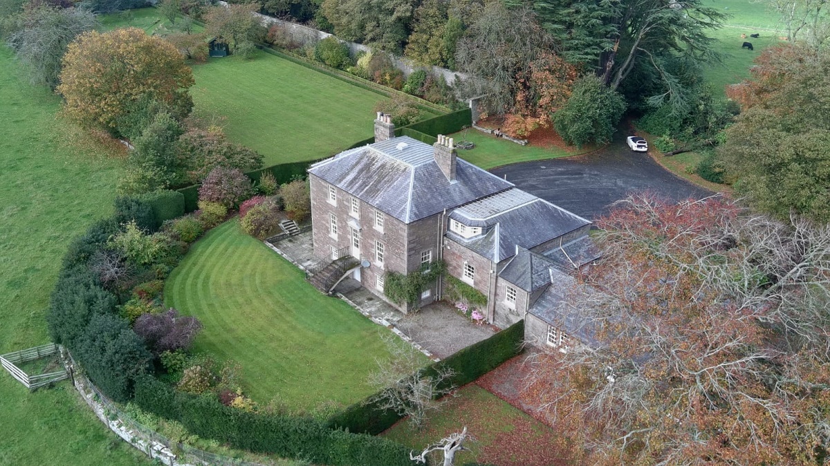 An elevated drone shot showing the large stone manor house nestled within expansive green lawns and mature trees. The property includes a paved driveway and is surrounded by a low stone wall and hedging.