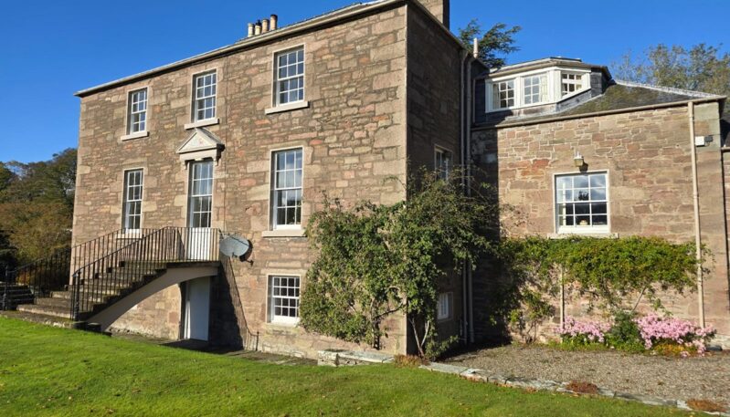 A ground-level view of the historic stone house under a clear blue sky. It highlights the multi-pane sash windows, a stone staircase leading to an elevated entrance, and well-maintained garden borders with pink flowers.