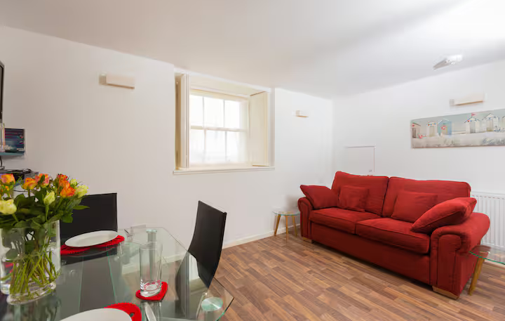 A bright, modern living and dining area featuring a vibrant red fabric sofa, a glass-top dining table with black chairs, and wood-effect flooring. A vase of orange roses sits on the table, and a small sash window with white shutters lets in natural light.