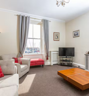 A cozy traditional lounge with high ceilings, featuring a large sash window overlooking a harbor with boat masts. The room includes a beige sofa with red tartan cushions, a large wooden coffee table, and a fireplace with a mantelpiece and mirror above it.