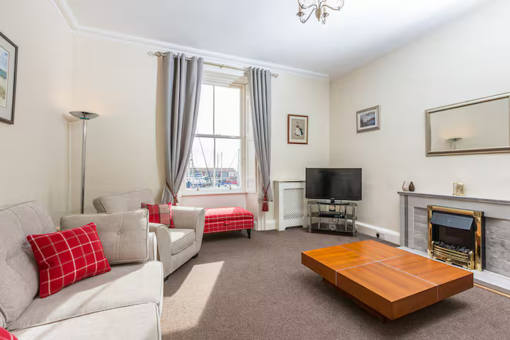 A cozy traditional lounge with high ceilings, featuring a large sash window overlooking a harbor with boat masts. The room includes a beige sofa with red tartan cushions, a large wooden coffee table, and a fireplace with a mantelpiece and mirror above it.