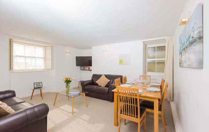 A spacious, open-plan living room with cream walls and carpet. It features two dark brown leather sofas, a glass coffee table, and a wooden dining set for four. Two sash windows provide light, and a flat-screen TV is mounted in the corner.