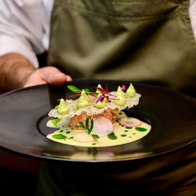 A chef in a green apron presents a meticulously plated appetizer on a large black dish. The dish features a base of tartare topped with a delicate, lacy crisp, dollops of green mousse, radish slices, microgreens, and a light creamy sauce