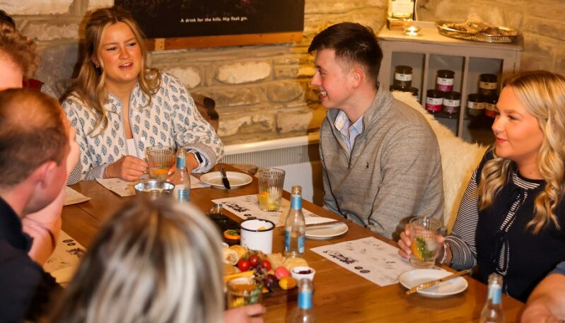 A group of friends sits around a long wooden table in a rustic room with exposed stone walls, enjoying a tasting experience. The table is filled with drinks, appetizers on wooden boards, and tasting menus, while a large landscape photograph of a stone bridge and heather hangs on the wall behind them.