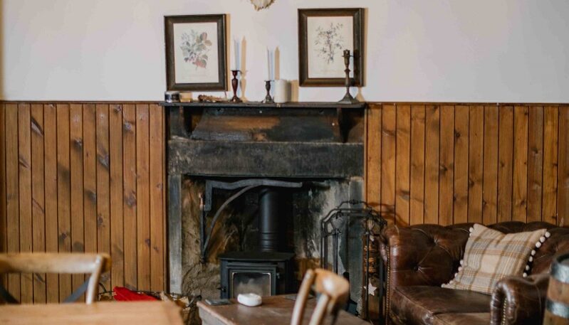 A cozy, rustic room featuring a stone fireplace with a wood-burning stove and a wood-paneled lower wall. Above the mantle, large deer antlers are mounted between two framed botanical prints, while a leather Chesterfield sofa, a wooden trunk coffee table, and a patterned rug complete the warm, lodge-style interior.