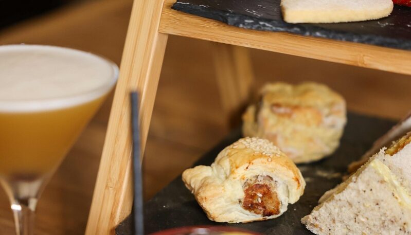 A close-up of a two-tiered wooden afternoon tea stand featuring slate platters. The top tier holds a pink meringue, a heart-shaped shortbread cookie, and a strawberry, while the bottom tier includes a sausage roll, a scone, and a finger sandwich. In the foreground, a foamy yellow cocktail in a coupe glass and a red berry-filled drink with a lemon slice sit on a wooden table.