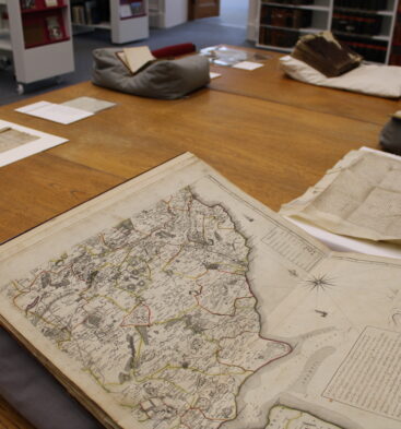 A display table featuring historic documents and an open antique map, with bookshelves visible in the background.