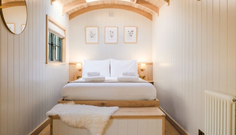 A cozy, narrow bedroom inside a shepherd's hut with white paneled walls and a curved wooden ceiling. A large bed with white linens and folded towels sits at the end of the room, positioned under three framed botanical prints and a small skylight. A white faux-fur rug rests on a wooden chest at the foot of the bed.