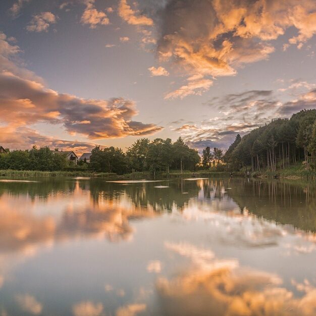 A peaceful landscape view of a still lake reflecting a dramatic sunset. The sky is filled with soft orange, pink, and purple clouds, which are mirrored perfectly in the water's surface. A dense line of dark green trees borders the right side of the lake, while a few small houses are visible among the foliage on the left.