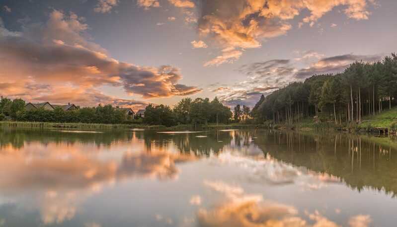 A peaceful landscape view of a still lake reflecting a dramatic sunset. The sky is filled with soft orange, pink, and purple clouds, which are mirrored perfectly in the water's surface. A dense line of dark green trees borders the right side of the lake, while a few small houses are visible among the foliage on the left.
