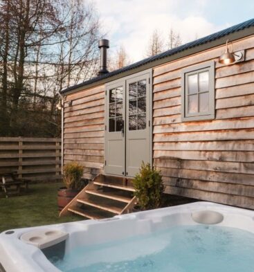 The exterior of a rustic wooden shepherd's hut with a curved corrugated roof and sage green double doors. In the foreground, a modern white hot tub is filled with bubbling water. The surrounding area includes a small patch of green grass, wooden steps leading into the hut, and a picnic table in the background near a wooden fence.