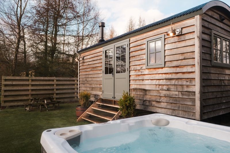 The exterior of a rustic wooden shepherd's hut with a curved corrugated roof and sage green double doors. In the foreground, a modern white hot tub is filled with bubbling water. The surrounding area includes a small patch of green grass, wooden steps leading into the hut, and a picnic table in the background near a wooden fence.