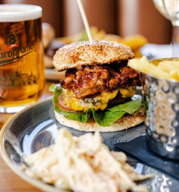 A close-up shot of a gourmet meal on a wooden table, featuring a large burger stacked with a beef patty, melted cheese, lettuce, tomato, and pulled pork on a sesame seed bun. The plate includes a side of coleslaw and a silver metal cup overflowing with french fries. In the background, a full pint glass of Staropramen beer sits next to a white napkin with silver cutlery.