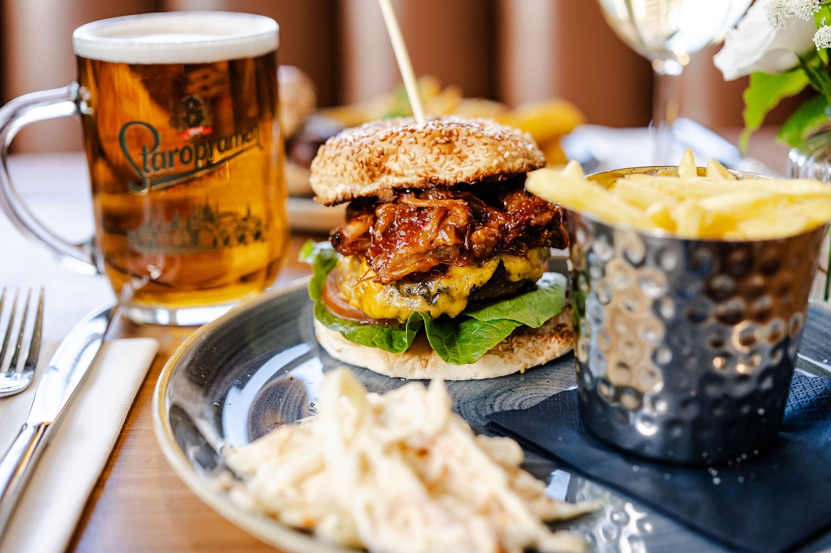 A close-up shot of a gourmet meal on a wooden table, featuring a large burger stacked with a beef patty, melted cheese, lettuce, tomato, and pulled pork on a sesame seed bun. The plate includes a side of coleslaw and a silver metal cup overflowing with french fries. In the background, a full pint glass of Staropramen beer sits next to a white napkin with silver cutlery.
