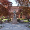 Statue standing in landscaped gardens outside Arbroath Library, a red sandstone civic building framed by mature trees, flowerbeds and paved pathways.