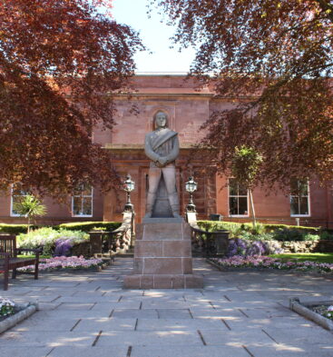 Statue standing in landscaped gardens outside Arbroath Library, a red sandstone civic building framed by mature trees, flowerbeds and paved pathways.