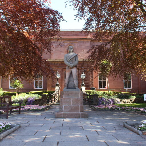 Statue standing in landscaped gardens outside Arbroath Library, a red sandstone civic building framed by mature trees, flowerbeds and paved pathways.