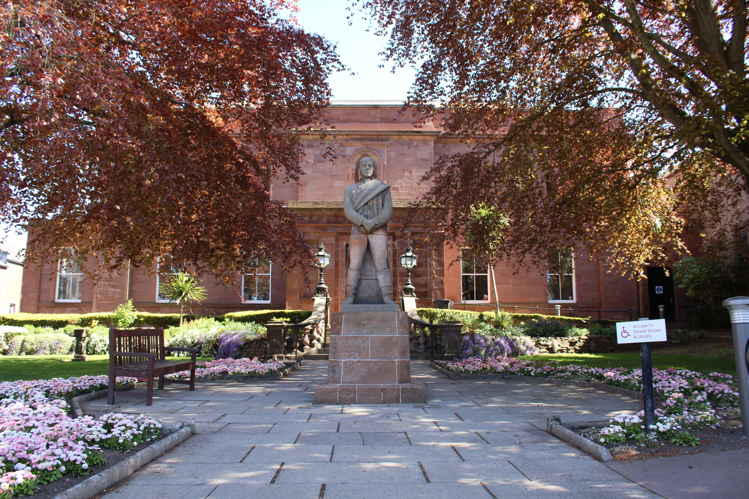 Statue standing in landscaped gardens outside Arbroath Library, a red sandstone civic building framed by mature trees, flowerbeds and paved pathways.