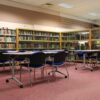 Interior of Forfar Library showing bookshelves, study tables and blue chairs arranged in a spacious reading area with carpeted flooring and overhead lighting.