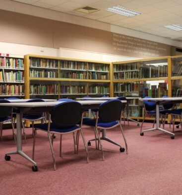 Interior of Forfar Library showing bookshelves, study tables and blue chairs arranged in a spacious reading area with carpeted flooring and overhead lighting.