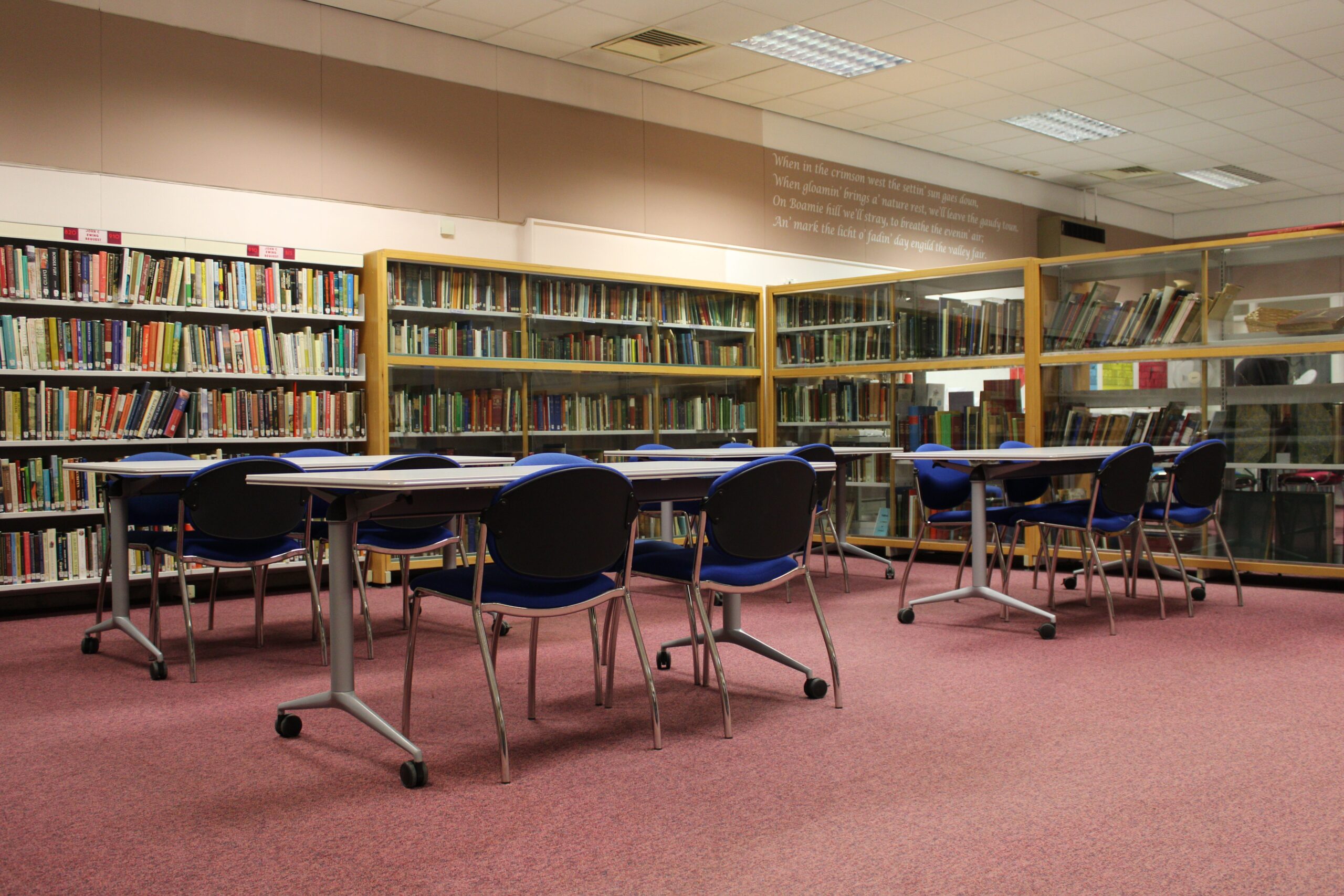 Interior of Forfar Library showing bookshelves, study tables and blue chairs arranged in a spacious reading area with carpeted flooring and overhead lighting.