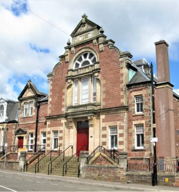 Historic red sandstone library building in Kirriemuir, featuring decorative Victorian architecture, arched windows, stone steps and a prominent central entrance facing the street.