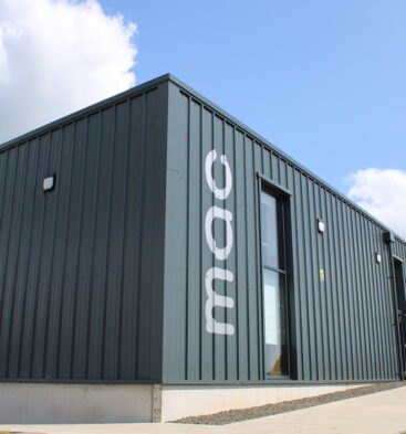 Modern Monifieth Library building with dark metal-clad exterior, large windows and accessible entrance pathways, set beneath a bright blue sky with scattered clouds.