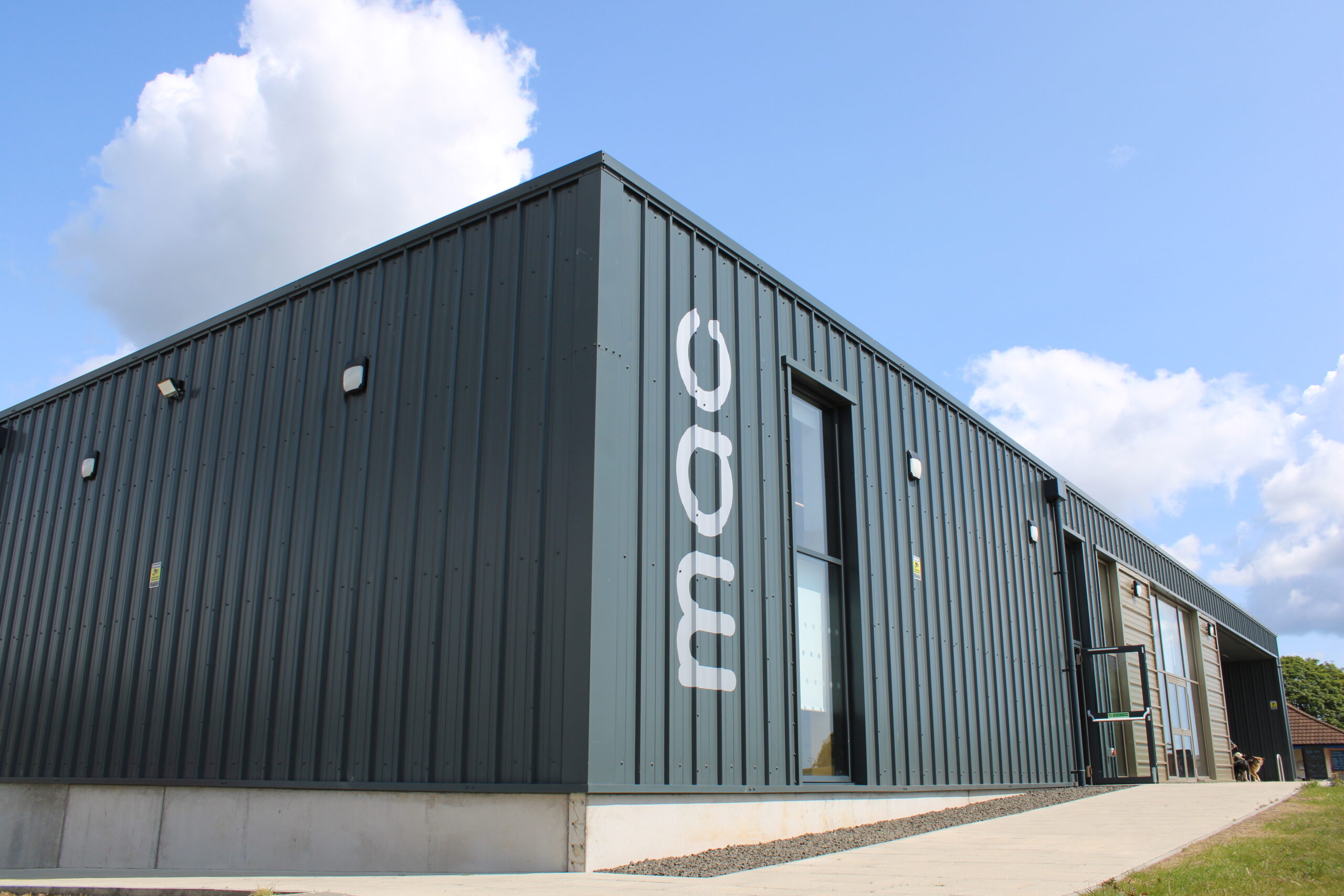 Modern Monifieth Library building with dark metal-clad exterior, large windows and accessible entrance pathways, set beneath a bright blue sky with scattered clouds.