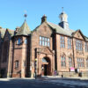 Historic red sandstone Montrose Library building featuring ornate Victorian architecture, large arched entrance, decorative stonework and tall windows, standing on a street corner beneath a clear blue sky.