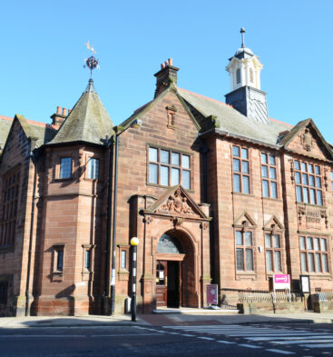 Historic red sandstone Montrose Library building featuring ornate Victorian architecture, large arched entrance, decorative stonework and tall windows, standing on a street corner beneath a clear blue sky.
