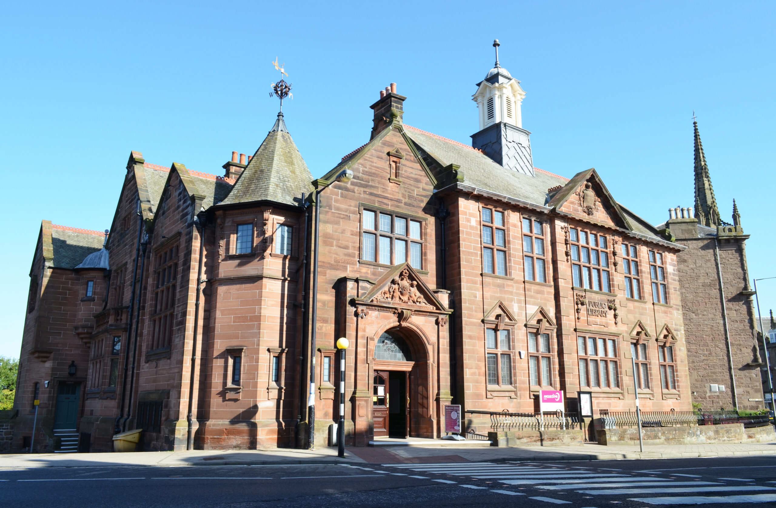 Historic red sandstone Montrose Library building featuring ornate Victorian architecture, large arched entrance, decorative stonework and tall windows, standing on a street corner beneath a clear blue sky.