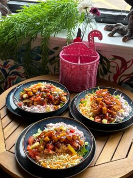 Three dark blue bowls filled with fried chicken rice bowls, topped with diced tomatoes, pineapple, dark sauce, shredded cheese, and creamy slaw, set on a wooden slatted table next to a bright pink woven flamingo utensil holder.