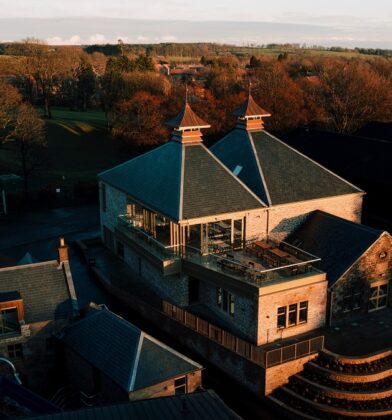 A high-angle sunset shot of the distillery, highlighting the modern glass-fronted visitor center balcony integrated with traditional stone architecture and the surrounding rolling hills.