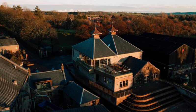 A high-angle sunset shot of the distillery, highlighting the modern glass-fronted visitor center balcony integrated with traditional stone architecture and the surrounding rolling hills.