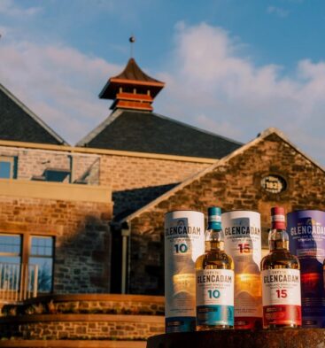 A close-up of three Glencadam Highland Single Malt Scotch Whisky bottles (10-year, 15-year, and Reserva de Jerez) and their decorative canisters, positioned in front of the distillery's stone buildings.